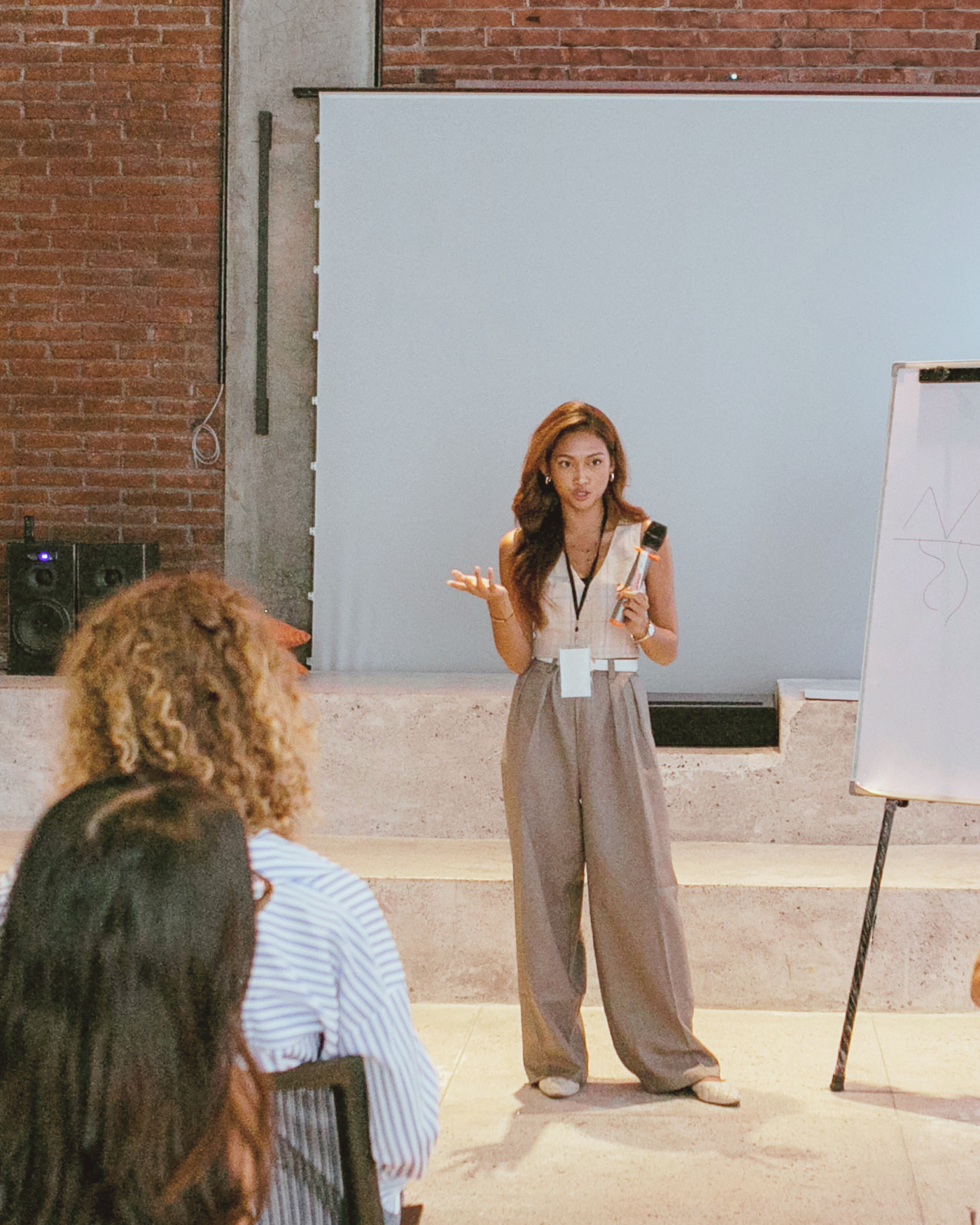 Woman presenting in a classroom setting with a whiteboard and brick wall.